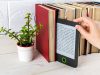 Mano che prende un e-reader su uno scaffale con libri cartacei e una piantina in vaso.