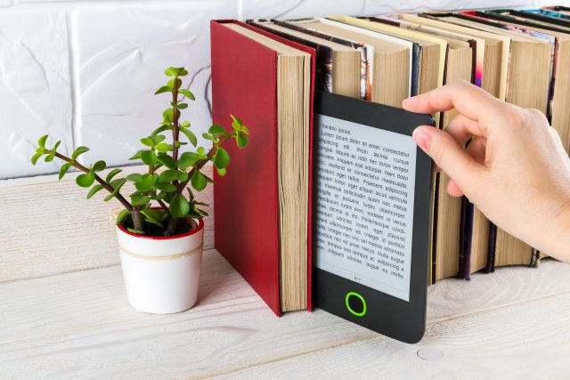 Mano che prende un e-reader su uno scaffale con libri cartacei e una piantina in vaso.